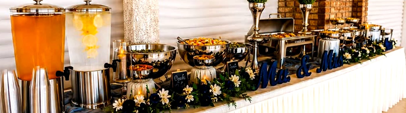 A wedding buffet set by White's Tasty Bites Catering — drink dispensers, chafing dishes, floral styling and a Mr & Mrs sign along a skirted table.
