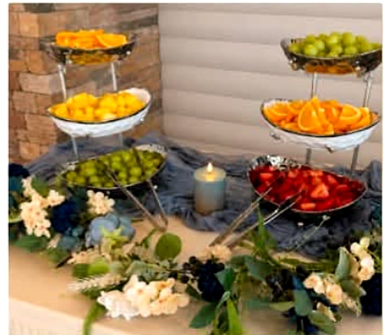A tiered fruit display of melon, grapes, orange slices and strawberries, dressed with flowers and candlelight.
